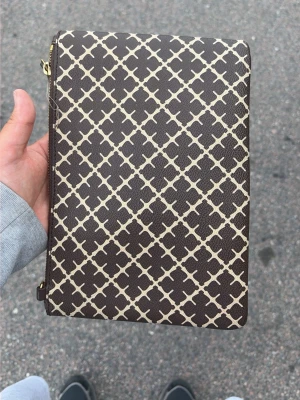 Brun mönstrad kuvertväska - Snygg brun kuvertväska med beige grafiskt mönster och guldfärgad dragkedja. Väskan har en rektangulär form och är tillverkad i ett tåligt material med ljus insida. Perfekt för att bära det viktigaste och ger en trendig touch till din outfit.