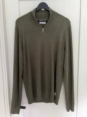 Olivgrön quarterzip - Stilren olivgrön tröja med quarterzip. Tröjan har långa ärmar och ribbade muddar vid ärmslut och nederkant. Perfekt för lager på lager och enkel att matcha med olika outfits.