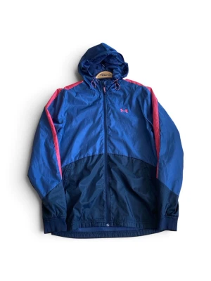 Blå vindjacka från Under Armour - Snygg blå vindjacka från Under Armour med rosa detaljer och logga på bröstet. Jackan har huva med dragsko, hel dragkedja framtill och elastiska muddar. Tillverkad i lätt och tåligt syntetmaterial, perfekt för blåsiga dagar.