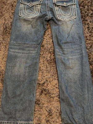 True Religion blå raka jeans - True Religion Joey Super T Jeans (W30) (äkta  Säljer ett par extremt snygga och ikoniska True Religion i modellen Joey Super T! Perfekt Y2K / streetwear-vibe med de klassiska, tjocka vita kontrastsömmarna och ikoniska locken på bakfickorna. Märke: True Religion (Made in USA) Modell: Joey Super T Storlek: W30 (Passar unisex! Sitter riktigt fräscht baggy/low waist på tjejer eller klassiskt på killar) Skick: Riktigt fint skick utan några problem