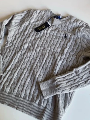 Grå kabelstickad tröja Polo Ralph Lauren - Snygg grå kabelstickad tröja från Polo Ralph Lauren med rund halsringning och ribbade muddar. Tröjan har den klassiska broderade loggan på bröstet och är tillverkad i mjukt material som passar perfekt till alla tillfällen.