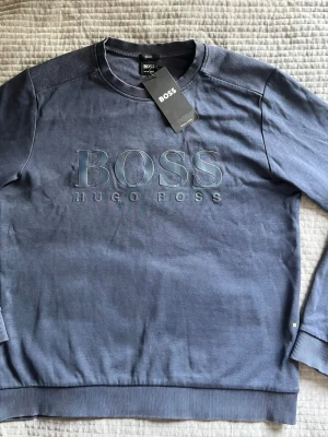 Blå sweatshirt från Hugo Boss - Snygg blå sweatshirt från Hugo Boss med stort broderat BOSS-tryck på bröstet. Tröjan har rund halsringning, ribbade muddar vid ärmslut och nederkant samt en avslappnad passform. Tillverkad i mjuk bomullsblandning för extra komfort. Tröjan sitter mer som en xs så lite förminskad!