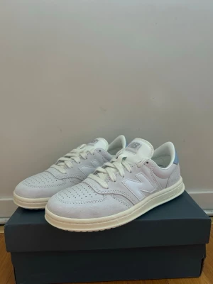 Vita New Balance sneakers i mocka - Snygga vita sneakers från New Balance i mocka och läder. Klassisk låg modell med perforerade detaljer och vit sula. Diskret gråblå detalj på hälen med New Balance-logga. Perfekta för en clean och stilren look.