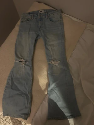 Ljusblå bootcut jeans med slitningar - Säljer ett par ljusblå bootcut jeans med slitningar över knäna. Jeansen har klassisk femficksmodell och är tillverkade i denim. Perfekta för en avslappnad och trendig look. Längden är short. 