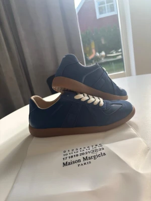 Blå Maison Margiela gats - Snygga blå Maison Margiela  sneakers med beige innersula och vita skosnören. Skorna har en klassisk siluett med gummisula och detaljer i både mocka och läder. Perfekta för dig som vill ha en stilren och trendig look.