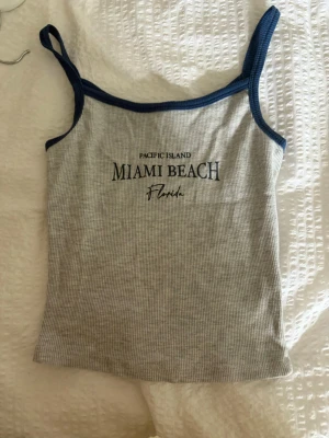 Grå ribbad linnetopp från Xside - Grå ribbad linnetopp från Xside med blå kantband och texten 'Miami Beach Florida' framtill. Toppen är ärmlös med smala axelband och har en slim passform, perfekt för varma dagar.