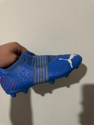 Blåa fotbollsskor från Puma - Säljer ett par blåa fotbollsskor från Puma med vita detaljer och röda inslag bak. Skorna har en strumpliknande passform, mönstrad ovandel och dobbar för spel på gräs. Perfekta för dig som vill ha bra grepp och kontroll på planen.