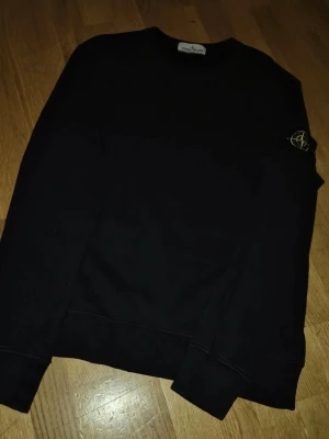 Svart sweatshirt från Stone Island - Svart sweatshirt från Stone Island med klassisk rund hals och ikonisk patch med logga på vänster ärm. Tröjan har ribbade muddar vid ärmslut och nederkant. Perfekt för en clean och stilren look.