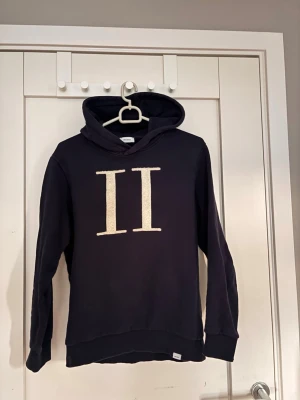Mörk blå hoodie med 2 streck  - Säljer en mörk blå hoodie från Les Deux. Den är i storlek 158-164 i barn. Hoodien har två styckna romerska ettor som är Les Deux logga. Ettorna är i ett luddigt material