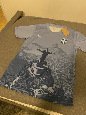 Brasil fotbollströja  - Snygg ljusblå fotbollströja från Nike med Brasilien-emblem och ikoniskt tryck av Kristusstatyn i Rio de Janeiro. Tröjan har rund hals och korta ärmar, perfekt för träning eller match.