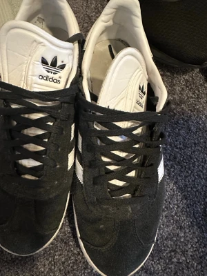 Svarta Adidas sneakers med vita detaljer - Klassiska svarta Adidas sneakers med vita ränder på sidorna och vit plös med Adidas-logga. Skorna har svart mockaliknande material på ovandelen och vita sulor. Låga i modellen och snörning framtill. Perfekta för en sportig och stilren look.