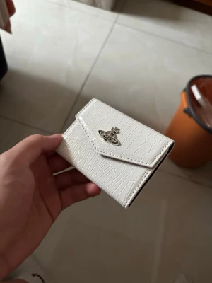 Vit plånbok med krokodilstruktur - Snygg vit plånbok i konstläder med krokodilpräglad yta och silverfärgad metallogo på framsidan. Plånboken har ett kuvertformat lock och kompakt design som gör den enkel att bära med sig. Perfekt för kort och kontanter.