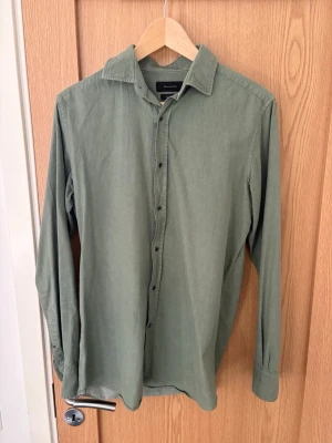 Grön långärmad skjorta i bomull - Snygg grön skjorta med klassisk krage och svarta knappar. Tillverkad i mjuk bomull och har lång ärm. Perfekt för en avslappnad men stilren look.