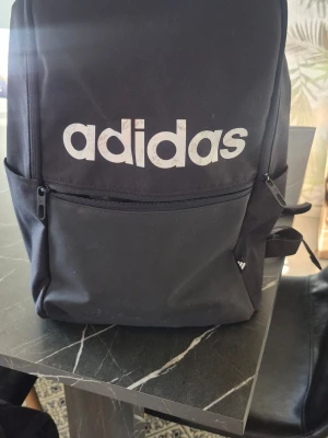 Svart ryggsäck från Adidas - Snygg svart ryggsäck från Adidas med stort vitt logotryck på framsidan. Ryggsäcken har en stor ytterficka med dragkedja, två sidofickor och justerbara axelband. Tillverkad i slitstarkt tygmaterial, perfekt för skolan eller träningen.