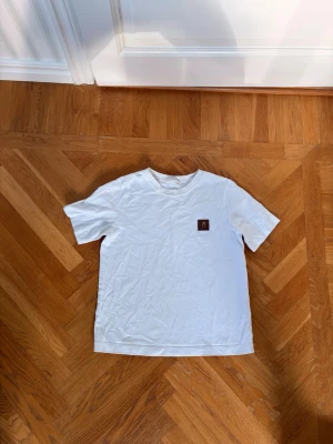 Vit t-shirt med patchdetalj - Vit t-shirt i bomull med rund hals och korta ärmar. På bröstet sitter en liten fyrkantig patch som ger en stilren touch. Perfekt basplagg för alla tillfällen och enkel att matcha med andra plagg.