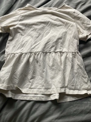 Vit volangblus från SAMSA Young - Söt vit blus från SAMSA Young i mjuk bomull. Toppen har korta ärmar och en volangdetalj nedtill som ger en avslappnad och trendig look. Perfekt att matcha med jeans eller kjol för en enkel och snygg outfit.