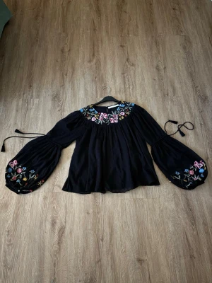 Svart blus med broderade blommor - Svart blus med vida, långa ärmar och färgglada blombroderier vid halsringningen och ärmsluten. Blusen har en lös passform och knytband vid ärmarna. Materialet ser ut att vara tunn och luftig tyg, perfekt för en trendig och bohemisk look. Kolla gärna mina andra annonser❗️