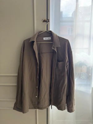 Beige overshirt från Samsøe Samsøe - Säljer en beige overshirt från Samsøe Samsøe. Skjortan har klassisk krage, bröstficka och knappar framtill. Perfekt att bära öppen över en t-shirt eller stängd för en clean look.