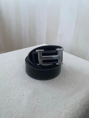 Svart skärp med stor H-spänne - Snyggt svart skärp i skinn med kontrasterande vita sömmar och ett stort, silverfärgat H-format spänne. Perfekt accessoar för att lyfta vilken outfit som helst och ger en clean, modern vibe.