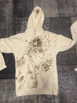 Vit hoodie från Gvy med tryck - Säljer en vit hoodie gvy med stor huva och dragsko. Framsidan har ett unikt brunt splash- och logotypmönster som ger en cool streetwear-vibe. Tillverkad i mjukt material och har ribbade muddar vid ärmar och nederkant.