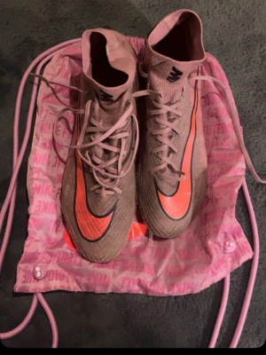 Nike rosa fotbollsskor med hög krage - Säljer ett par snygga fotbollsskor från Nike i rosa med orange detaljer och hög krage för extra stöd. Skorna har klassisk snörning och en modern, strömlinjeformad design. Perfekta för dig som vill sticka ut på planen!