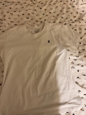 Vit t-shirt  - Klassisk vit t-shirt med den ikoniska broderade loggan på bröstet. T-shirten har rund hals och korta ärmar, tillverkad i mjuk bomull för en skön känsla. Perfekt basplagg till garderoben.