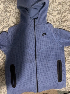 Blå Nike hoodie med dragkedja - Säljer en blå hoodie från Nike med hel dragkedja framtill och två svarta dragkedjeförsedda fickor. Tröjan har en strukturerad yta och en stor huva. Nike-loggan syns på bröstet. Perfekt för dig som gillar sportig och avslappnad stil.