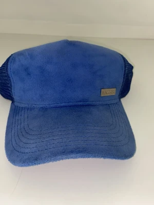 Blå truckerkeps från Maggiore - Snygg blå truckerkeps från Maggiore med mjuk, sammetsliknande framsida och mesh på sidorna och bak. Justerbar snapback för perfekt passform. Metallogo framtill ger en clean och modern look. Perfekt för dig som vill sticka ut med stil.