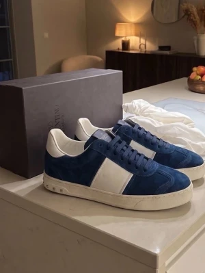 Blå och vita sneakers från Valentino - Snygga sneakers i blå mocka och vitt läder från Valentino. Skorna har en klassisk låg silhuett, vita detaljer på sidan och häl, samt vita snören och sula. Perfekta för dig som vill ha en stilren och trendig look.