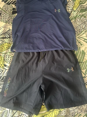 Under Armour träningsset - Säljer ett träningsset från Under Armour med en blå t-shirt och svarta shorts. T-shirten har rund hals, tryckt logga och texten UNDER ARMOUR i regnbågsfärg. Shortsen har dragsko i midjan, logga och texten ARMOUR på benet. Perfekt för gym eller löpning.