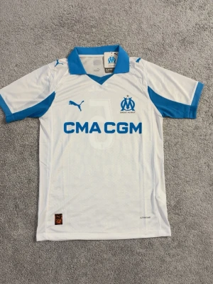 Marseille matchtröja Puma vit/blå - Säljer en vit och blå Olympique de Marseille matchtröja från Puma med tryck 'CMA CGM' och nummer 5 samt 'JUL' på ryggen. Tröjan har korta ärmar, blå krage och detaljer, samt klubbmärke och Puma-logga på bröstet. Perfekt för fotbollsfans!