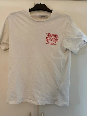Vit Replay t-shirt med röd print (äkta) - Säljer en vit t-shirt från Replay med röd textprint både fram och bak. Klassisk rund halsringning och korta ärmar. Materialet är mjuk bomull som känns skönt mot huden. Perfekt för dig som gillar enkel men snygg stil.