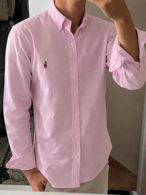 Ljusrosa skjorta från Polo Ralph Lauren - Klassisk ljusrosa skjorta från Polo Ralph Lauren med button-down krage och broderad logga på bröstet. Skjortan har lång ärm, normal passform och är tillverkad i mjuk bomull. Perfekt för en clean och stilren look. M men sitter som S