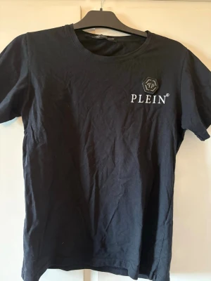 Philipp plein T-shirt  - Snygg svart t-shirt från Plein med vit logga och emblem på bröstet. Klassisk rund halsringning och korta ärmar. Tillverkad i mjukt bomullstyg som känns skönt mot huden. Perfekt till jeans eller shorts för en clean look.