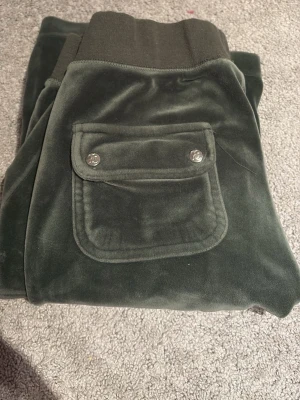 Mörkgröna velour mjukisbyxor - Mörkgröna mjukisbyxor i mjuk velour med bred mudd i midjan och en ficka med lock och silverfärgade knappar bak. Perfekta för chill dagar och har en riktigt skön känsla. Snygg detalj med fickan och klassisk avslappnad passform. Saknar snören men arnas är dom som nya säljer bara för att dom är förstora för mig och använder inte längre. Pris kan diskuteras 