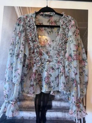 Blommig volangblus från Zara - Zara blus i tunn, ljusblå mesh med blommönster i rosa, lila och grönt. Djup V-ringning med volangdetaljer och dekorativa knappar framtill. Vida, långa ärmar med resår och volang vid ärmslut. 