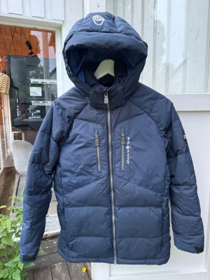 Mörkblå dunjacka med huva - Snygg mörkblå dunjacka med stor huva och två vertikala bröstfickor med dragkedja. Jackan har quiltad design och logga på både huva och bröst. Perfekt för kalla vinterdagar och riktigt varm tack vare dunfyllningen.