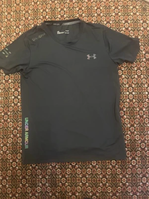 Svart träningströja Under Armour - Snygg svart träningströja från Under Armour med korta ärmar och rund hals. Loggan syns på bröstet och texten UNDER ARMOUR i regnbågsfärger längs sidan. Tillverkad i ett snabbtorkande funktionsmaterial som passar perfekt till träning. Storlek M passar S