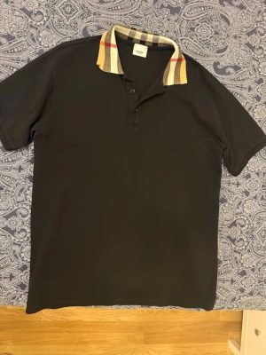 Svart pikétröja från Burberry - Säljer en svart pikétröja från Burberry med klassisk rutig krage i beige, svart, vit och röd. Tröjan har korta ärmar och knappar framtill. Perfekt för dig som vill ha en stilren och tidlös look.