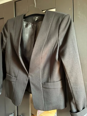 Svart figursydd kavaj - Stilren svart kavaj med figurnära passform och klassisk krage. Kavajen har två fickor framtill och är helfodrad med slät insida. Perfekt för en dressad look och enkel att matcha med olika outfits.