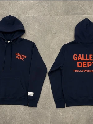 Mörkblå hoodie från Gallery Dept - Snygg mörkblå hoodie från Gallery Dept med huva och känguruficka. Tryck i rött framtill och stort tryck på ryggen med texten 'GALLERY DEPT. HOLLYWOOD, CA.'. Tillverkad i mjukt bomullsmaterial, perfekt för en avslappnad streetwear-look.