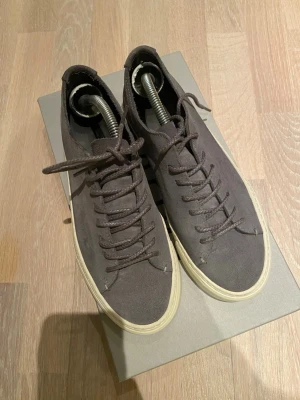 Mörkgrå mockasneakers med vit sula - Stilrena sneakers i mörkgrå mocka med klassisk vit sula. Skorna har rund tå, matchande mörkgrå skosnören och diskret design utan synliga loggor. Perfekta för dig som gillar minimalistisk och clean stil.