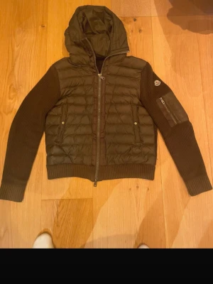 Brun pufferjacka från Moncler - Snygg brun pufferjacka från Moncler med stickade ärmar och huva. Jackan har quiltad front, dragkedja och Moncler-logga på ärmen. Perfekt för kalla vinterdagar och har en modern, sportig look. Materialmix av dun och stickat ger både värme och stil! Modellen är i sig vanligts lite kortare men sitter perfekt!
