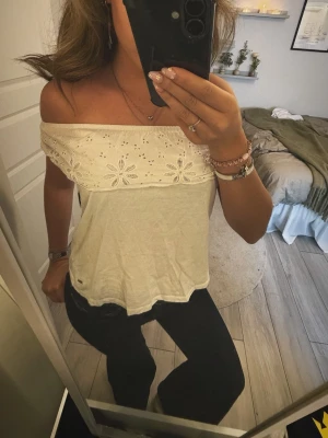 Vit offshoulder topp med spetsdetaljer - Säljer en söt vit offshoulder topp med bred spetskant och broderade blommor. Toppen är ärmlös och har en luftig passform, perfekt för varma dagar. 