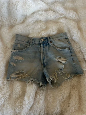 Ljusa slitna jeansshorts med hög midja - Säljer ett par ljusblå jeansshorts med hög midja och flera slitningar och hål för en trendig, avslappnad look. Shortsen har råa kanter, klassiska fem fickor och knäppning med knappar framtill. Perfekta för dig som gillar en edgy stil.