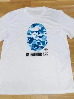 Vit BAPE t-shirt med blått camomönster - Säljer en vit t-shirt från By Bathing Ape med det klassiska blå camomönstret och BAPE-loggan på bröstet. T-shirten har rund hals och korta ärmar, perfekt för dig som gillar streetwear och vill sticka ut.