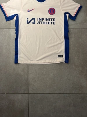 Chelsea FC vit matchtröja Nike - Chelsea FC:s officiella vita matchtröja från Nike med blåa sidopaneler och röda detaljer. Tröjan har klubbmärke och Nike-logga på bröstet samt sponsortryck 'INFINITE ATHLETE' framtill. Tillverkad i lätt och ventilerande funktionsmaterial.
