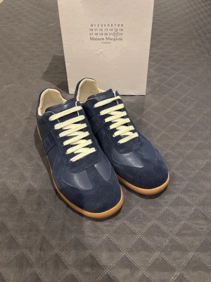 Blå Maison Margiela sneakers - Snygga blå sneakers från Maison Margiela med ovandel i mocka och läder, vita snören och klassisk gummisula. Skorna har en låg silhuett och stilren design som passar till många olika outfits.