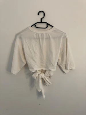 Vit croppad topp med justerbar rosett vid ryggen - Säljer en vit croppad topp med justerbar rosett vid ryggen och trekvartsärm. Toppen har en enkel och clean design med rund halsringning. Perfekt för en avslappnad och trendig look.