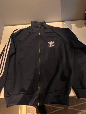 Marinblå Adidas track jacket - Klassisk marinblå Adidas track jacket med vita ränder längs ärmarna och dragkedja framtill. Tröjan har hög krage, två fickor och Adidas Trefoil-logga på bröstet och ryggen. Perfekt för en sportig och avslappnad stil.
